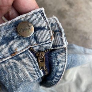 May include: Close-up of a light blue denim jeans waistband. The jeans feature a brass-colored button with the brand name "Wrangler" and a brass zipper. The denim has a faded, worn appearance.