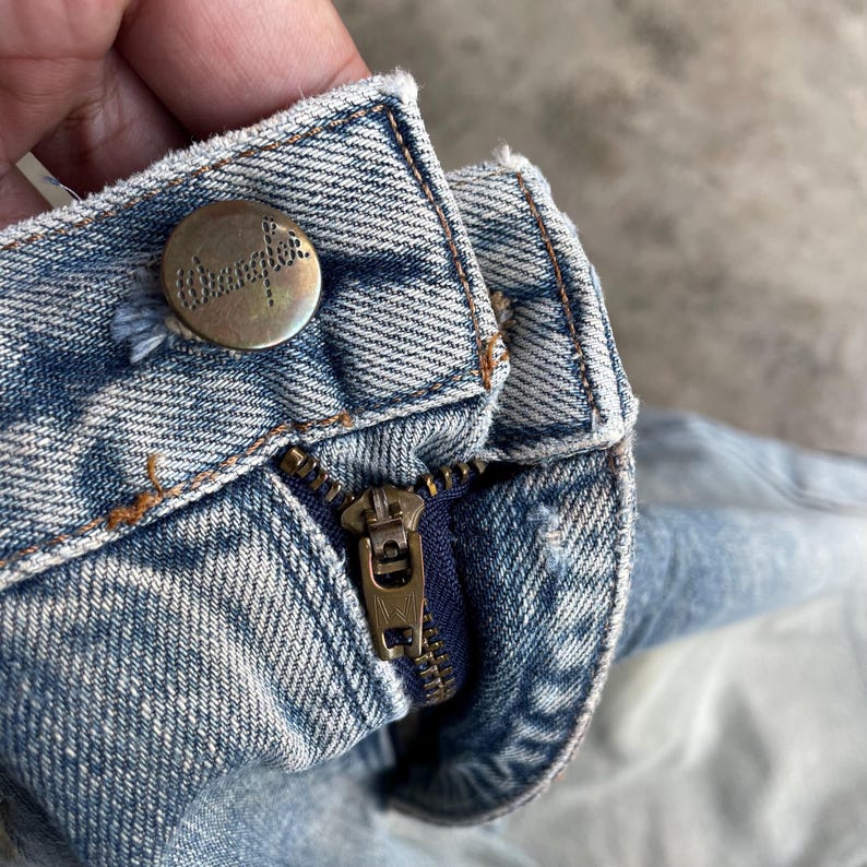 May include: Close-up of a light blue denim jeans waistband. The jeans feature a brass-colored button with the brand name "Wrangler" and a brass zipper. The denim has a faded, worn appearance.