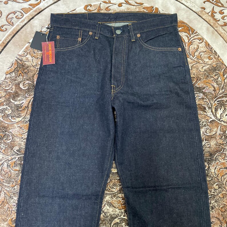 May include: Dark wash denim jeans with a classic five-pocket design. The jeans feature a button and zipper closure, with visible stitching in a contrasting color. A tag with the text "JOE McCOY" is attached.