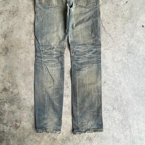 May include: Faded blue denim jeans with distressed details. The jeans have a classic five-pocket design, a button and zipper closure, and a worn appearance. The denim shows signs of wear and tear, with faded areas and subtle distressing.