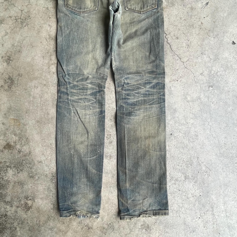 May include: Faded blue denim jeans with distressed details. The jeans have a classic five-pocket design, a button and zipper closure, and a worn appearance. The denim shows signs of wear and tear, with faded areas and subtle distressing.