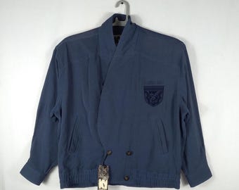 May include: A navy blue jacket with a distinctive wrap-style collar and two rows of buttons. The jacket has a patch with text and a design on the left chest. It has long sleeves and a ribbed hem. A tag hangs from the jacket.