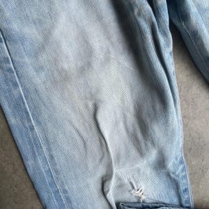 May include: Light blue denim jeans with a faded wash and distressed details. The jeans have a classic five-pocket design and a relaxed fit. The denim is slightly worn, with a small tear near the knee.