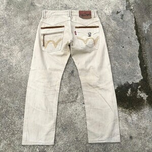May include: Beige denim jeans with brown leather accents and embroidered details. The back pockets have a unique W-shaped design.