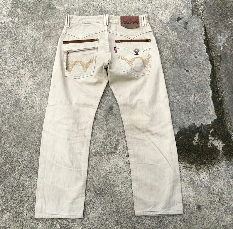 May include: Beige denim jeans with brown leather accents and embroidered details. The back pockets have a unique W-shaped design.