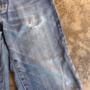 Pu&ograve; includere: Primo piano di jeans di denim blu strappati. I jeans mostrano segni di usura, con aree sbiadite e un piccolo strappo. Il tessuto ha un aspetto strutturato, con cuciture visibili lungo le cuciture.
