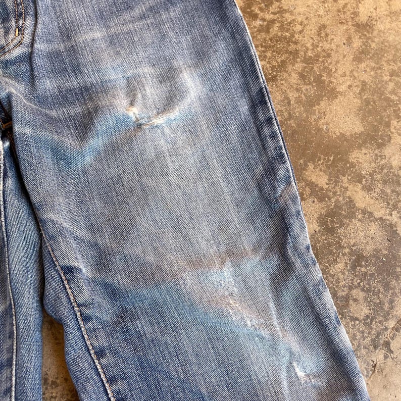 Pu&ograve; includere: Primo piano di jeans di denim blu strappati. I jeans mostrano segni di usura, con aree sbiadite e un piccolo strappo. Il tessuto ha un aspetto strutturato, con cuciture visibili lungo le cuciture.