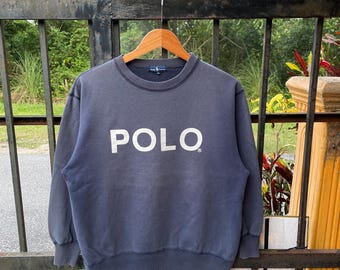 May include: A navy blue sweatshirt with the word "POLO" in white block letters. The crew neck sweatshirt is displayed on a wooden hanger. The background features greenery and a metal fence.