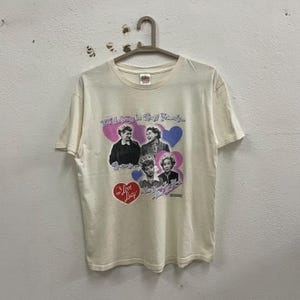 May include: A white t-shirt with a graphic print of the cast of I Love Lucy. The graphic features the four main characters, Lucille Ball, Vivian Vance, Desi Arnaz, and William Frawley, with the text "We'll always be best friends..." and "I Love Lucy".
