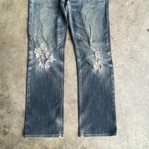 K&ouml;nnte beinhalten: Verblasste blaue Jeans mit abgenutzten Details, einschlie&szlig;lich zerrissener Patches an den Knien. Die Jeans haben ein klassisches F&uuml;nf-Taschen-Design und einen geraden Beinverlauf. Der Denim weist Gebrauchsspuren und Ausbleichungen auf.