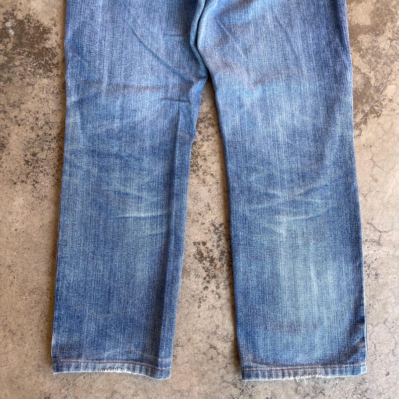 Pu&ograve; includere: Jeans di denim blu chiaro con un lavaggio sbiadito. I jeans hanno un classico design a cinque tasche e un taglio dritto. Il tessuto mostra segni di usura, aggiungendo all'estetica vintage.