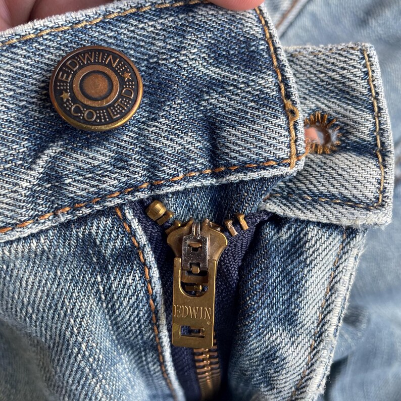 May include: Close-up of light blue denim jeans. The jeans have a brass button with "EDWIN & CO." and a brass zipper with "EDWIN" engraved. The stitching is tan. The jeans are a classic style.