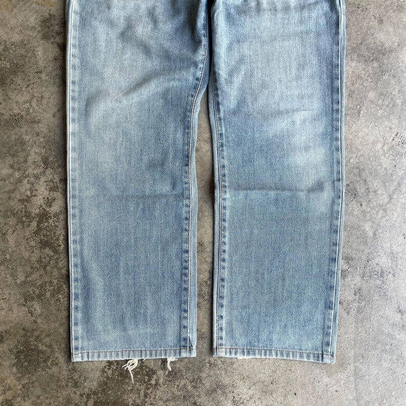 May include: Light blue denim jeans with a straight leg cut. The jeans have a classic five-pocket design and a faded wash. The bottom hem of the jeans is slightly frayed.
