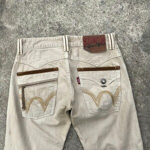 May include: Beige denim jeans with brown leather accents and a zippered pocket on the back. The jeans have a brown leather patch with the text "EDWIN E-clusive Vintage" on the back waistband.