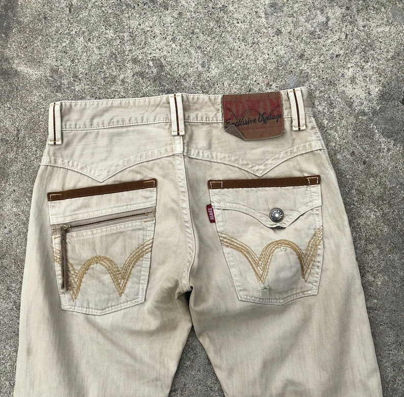 May include: Beige denim jeans with brown leather accents and a zippered pocket on the back. The jeans have a brown leather patch with the text "EDWIN E-clusive Vintage" on the back waistband.