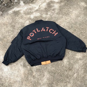 May include: Black bomber jacket featuring the word "POTLATCH" in orange lettering on the back, with "80-East" underneath. The jacket has a ribbed collar, cuffs, and waistband. A classic, casual outerwear piece.