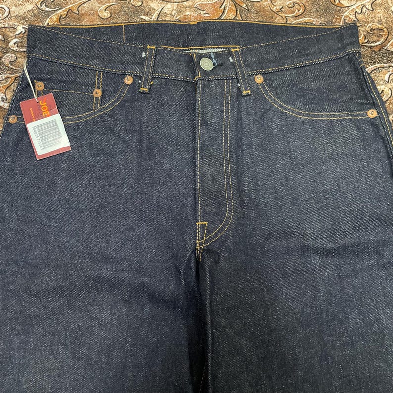 May include: Dark wash denim jeans with a classic five-pocket design. The jeans feature yellow stitching, copper rivets, and a button closure. A red tag with the brand name "JOE" is attached to the front pocket.