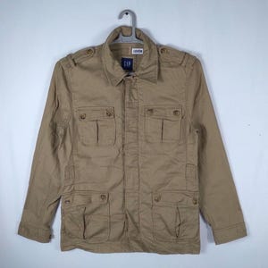 Puede incluir: Una chaqueta utilitaria de color tostado con mangas largas, cuello, cierre frontal de botones y cuatro bolsillos delanteros con solapas abotonadas. La chaqueta incluye charreteras en los hombros y una etiqueta azul de Gap en el cuello.