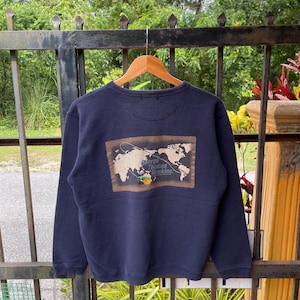 Peut inclure: Sweat-shirt bleu marine avec un motif de carte du monde et le texte "George Thomas Hawkins". Le motif est dans les tons marron et beige. Le sweat-shirt est suspendu à un cintre en bois.