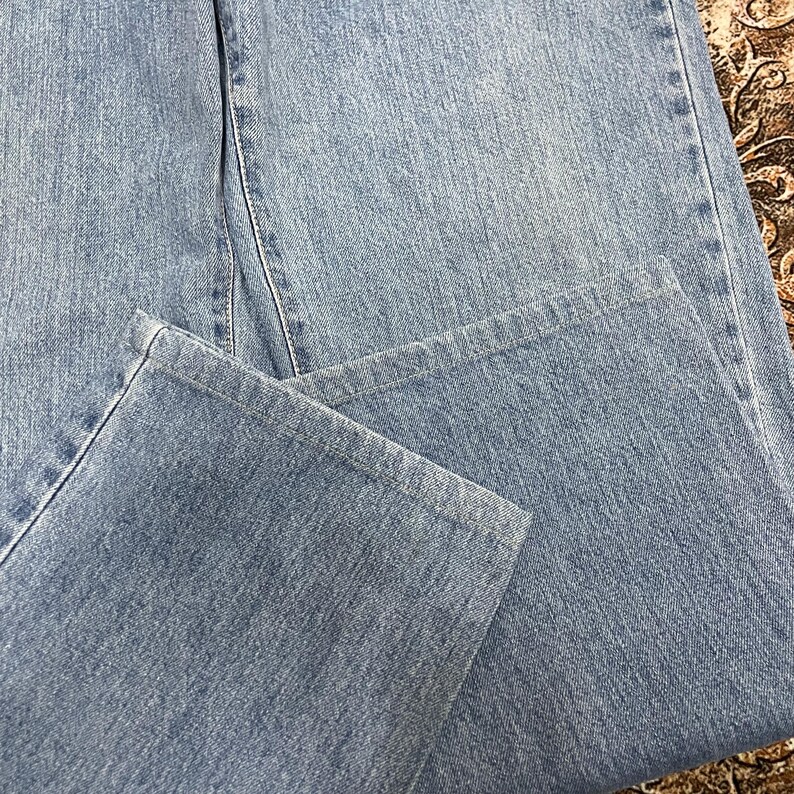 May include: A pair of light blue denim jeans featuring a classic straight-leg cut with visible stitching along the seams and hem. The fabric displays a consistent medium-wash texture with subtle variations in the blue tone across the surface.