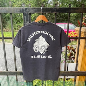 May include: Dark grey t-shirt with a graphic on the back. The graphic shows a cobra on a motorcycle, with the text "DUAL SERPENTINE FORCE" and "U.S. AIR BASE MC." The t-shirt is hanging on a wooden hanger.