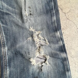 K&ouml;nnte beinhalten: Nahaufnahme einer Jeans aus blauem Denim mit einem gro&szlig;en, ausgefransten Loch. Der Stoff weist Abnutzungserscheinungen auf, mit helleren Flecken und dunkleren Flecken. Die Jeans hat sichtbare N&auml;hte und ein strukturiertes Aussehen.