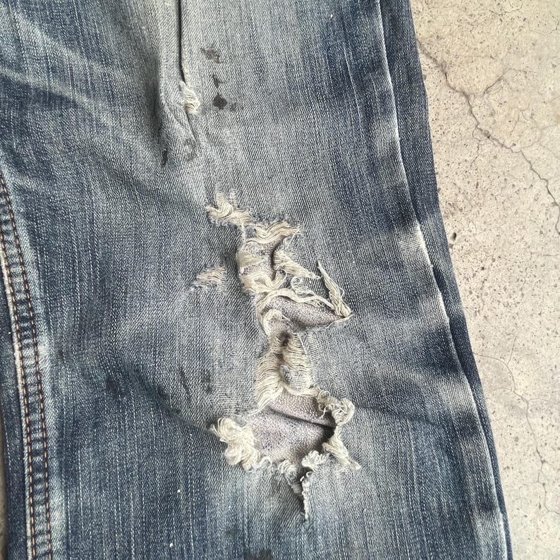 K&ouml;nnte beinhalten: Nahaufnahme einer Jeans aus blauem Denim mit einem gro&szlig;en, ausgefransten Loch. Der Stoff weist Abnutzungserscheinungen auf, mit helleren Flecken und dunkleren Flecken. Die Jeans hat sichtbare N&auml;hte und ein strukturiertes Aussehen.