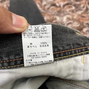 May include: Close-up of a tag from a pair of black denim jeans. The tag has Japanese text and symbols, including washing instructions and fabric composition. The fabric is 100% cotton with a synthetic leather label. Made in China.