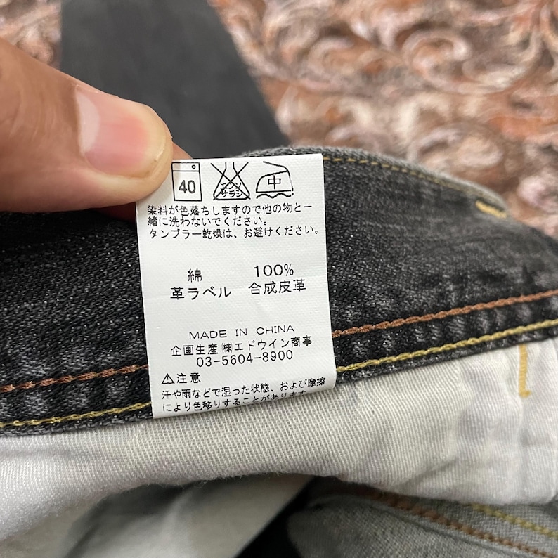 May include: Close-up of a tag from a pair of black denim jeans. The tag has Japanese text and symbols, including washing instructions and fabric composition. The fabric is 100% cotton with a synthetic leather label. Made in China.