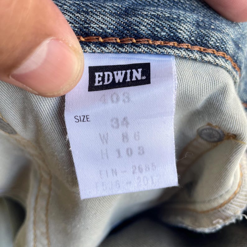 May include: Close-up of a denim jeans label with the brand name "EDWIN" in white on a black rectangle. The label shows size information: 34 waist, 86 cm length, and 103 cm height. The jeans are blue with visible stitching.