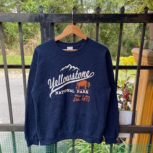 Może przedstawiać: Niebieska bluza z długim rękawem z białym nadrukiem graficznym logo Parku Narodowego Yellowstone. Logo przedstawia pasmo górskie, bizona i tekst "Yellowstone National Park, Oldest & Best, Est. 1872."