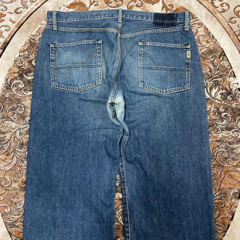 May include: A pair of blue denim jeans featuring a classic five-pocket design, a medium wash finish, and a dark rectangular patch on the back waistband. The back pockets include decorative stitching, and the fabric shows signs of wear.