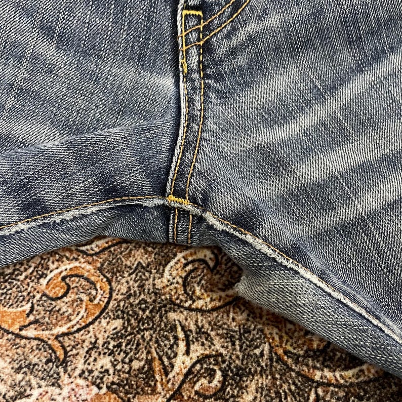 May include: A pair of blue denim jeans featuring a faded wash and visible texture. The crotch area shows reinforced seams stitched with thick, golden-yellow thread, highlighting the construction details of the fabric panels and the denim weave.