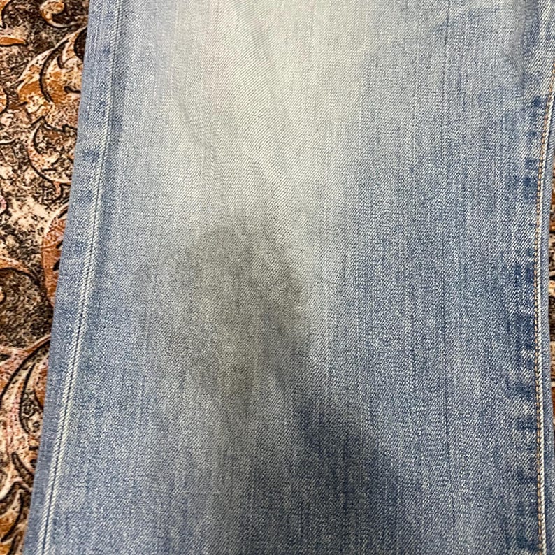 May include: Close-up of light blue denim jeans. The jeans have a faded appearance with subtle variations in color. The fabric shows a textured weave, and the seams are visible along the edges. The jeans are slightly worn, suggesting regular use.