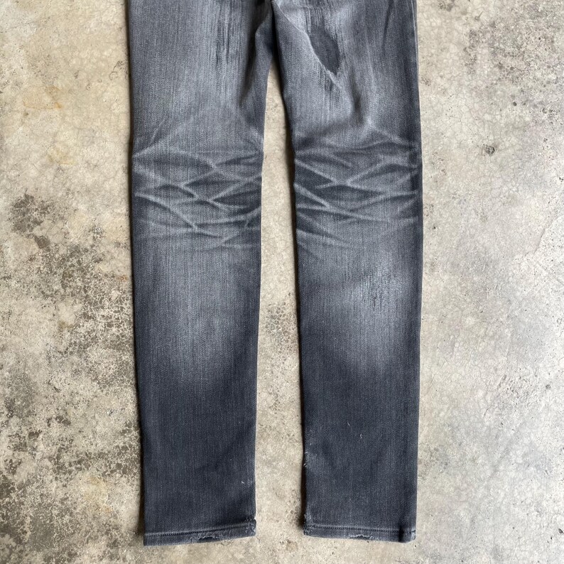 May include: A pair of faded black denim jeans. The jeans have a straight leg cut and show wear marks on the knees. The fabric has a slightly distressed appearance, with subtle variations in color throughout the garment.