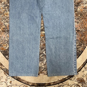May include: A pair of light blue denim jeans featuring a straight-leg cut with visible vertical stitching along the outer seams. The fabric displays a consistent medium-wash texture throughout the legs, ending in a clean, straight-hemmed finish.