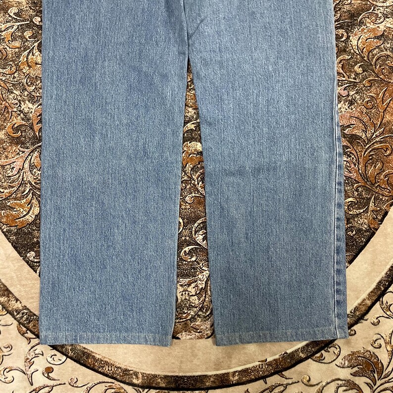 May include: A pair of light blue denim jeans featuring a straight-leg cut with visible vertical stitching along the outer seams. The fabric displays a consistent medium-wash texture throughout the legs, ending in a clean, straight-hemmed finish.