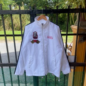 May include: White zip-up jacket with black stripes down the sleeves. Features a skull patch with a checkered flag and the brand name "Von Dutch" in red script. The jacket has a high collar and two front pockets.