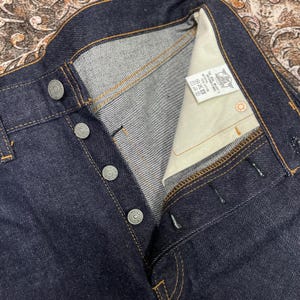 May include: Close-up of dark blue denim jeans with a button fly. The jeans have silver buttons and visible yellow stitching. A white tag with text is visible inside the waistband. The jeans are new and unwashed.