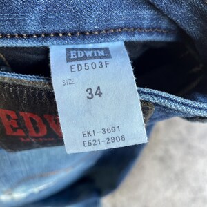 Op de afbeelding: Close-up van een blauw denim jeanslabel met de merknaam "Edwin" en het modelnummer "ED503F". Het label geeft maat 34 aan. Extra codes "EK1-3691" en "E521-2806" zijn ook zichtbaar. De jeans is gemaakt van blauw denim.