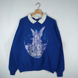 May include: A blue sweatshirt with a white collar and a graphic print of an angel with wings. The angel is surrounded by a blue and white design.
