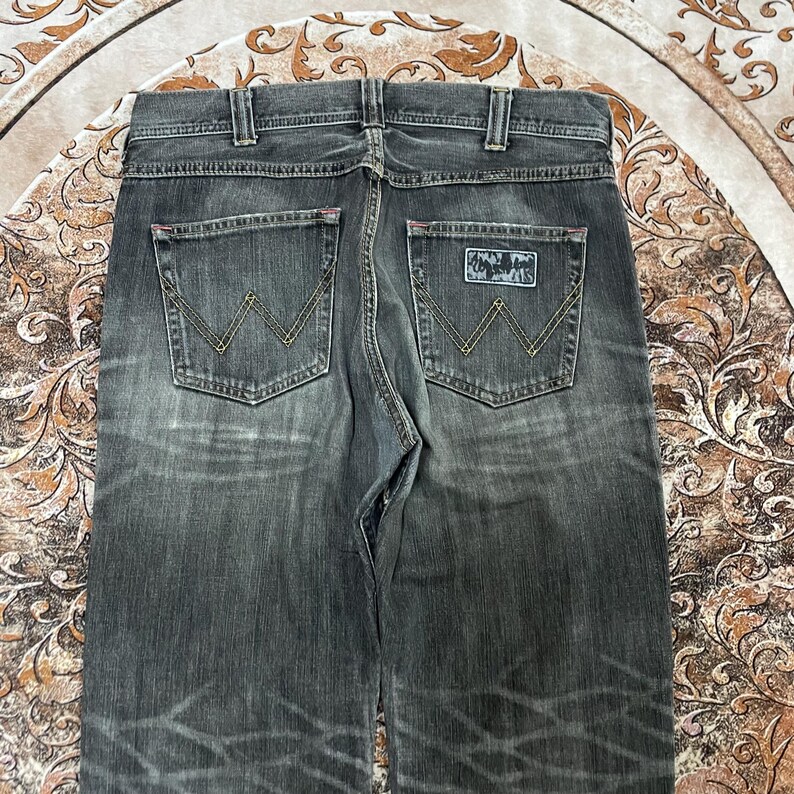 May include: Dark gray denim jeans with a faded wash effect. The back pockets feature a yellow stitched 'W' design, and a small patch with text is visible on the right pocket. The jeans have a classic five-pocket design.