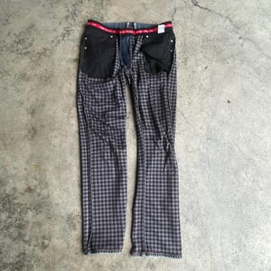 K&ouml;nnte beinhalten: Eine Jeanshose mit grau-schwarzem Karomuster und rotem Bund. Die Jeans liegt flach, wodurch die Innentaschen und der Rei&szlig;verschluss sichtbar werden. Der Stoff hat ein ausgepr&auml;gtes Karomuster.