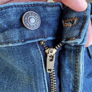 Op de afbeelding: Close-up van een blauwe spijkerbroek met een metalen knoop en rits. De knoop is voorzien van de merknaam "EDWIN". De rits is goudkleurig en heeft ook de merknaam "EDWIN" erop. De jeans heeft zichtbare stiksels.