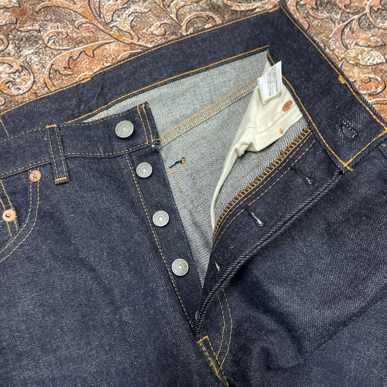 May include: Close-up of dark blue denim jeans with button fly and metal rivets. The jeans have a classic five-pocket design with visible stitching and a partially unbuttoned fly. The fabric appears to be a sturdy, raw denim.