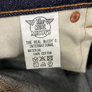 May include: Close-up of a label from The Real McCoy's jeans. The label is white with black text, including the brand name and "100% COTTON." A copper-colored button is visible.