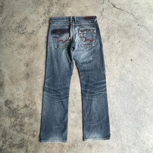 K&ouml;nnte beinhalten: Verblasste blaue Jeans mit geradem Bein. Zu den Merkmalen geh&ouml;ren dunkelblaue Taschen mit roten N&auml;hten und ein rotes Logo-Patch auf der R&uuml;ckseite. Die Jeans haben einen abgenutzten, verwitterten Look.