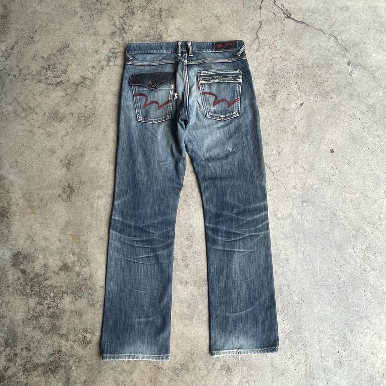 K&ouml;nnte beinhalten: Verblasste blaue Jeans mit geradem Bein. Zu den Merkmalen geh&ouml;ren dunkelblaue Taschen mit roten N&auml;hten und ein rotes Logo-Patch auf der R&uuml;ckseite. Die Jeans haben einen abgenutzten, verwitterten Look.