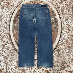 May include: A pair of blue denim jeans shown from the back, featuring a medium wash, two back pockets with horizontal stitching details, a dark rectangular patch on the waistband, and visible wear and fading at the hems and crotch area.