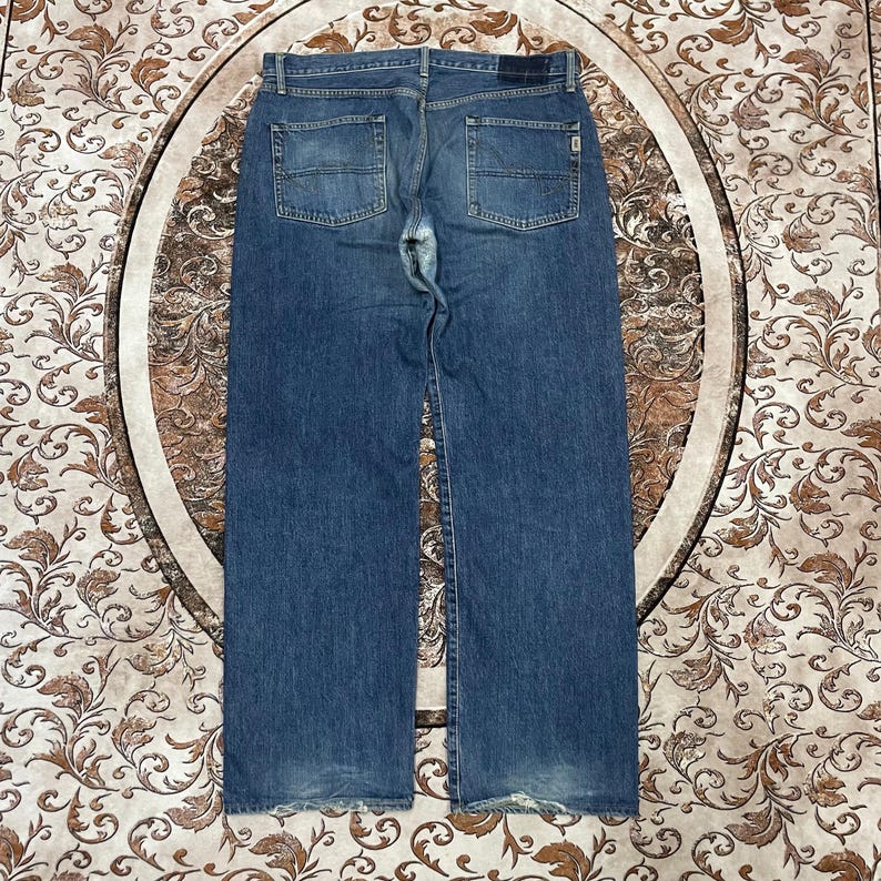 May include: A pair of blue denim jeans shown from the back, featuring a medium wash, two back pockets with horizontal stitching details, a dark rectangular patch on the waistband, and visible wear and fading at the hems and crotch area.